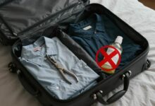 Uma mala de viagem aberta sobre uma cama, com roupas e objetos sendo arrumados, e alguns itens proibidos, como uma tesoura grande e um frasco de líquido maior que 100ml, com um X vermelho sobre eles.
