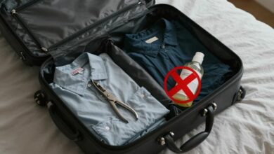 Uma mala de viagem aberta sobre uma cama, com roupas e objetos sendo arrumados, e alguns itens proibidos, como uma tesoura grande e um frasco de líquido maior que 100ml, com um X vermelho sobre eles.