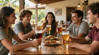Um grupo de amigos brindando com diferentes tipos de copos de cerveja artesanal em uma cervejaria rústica e ensolarada.